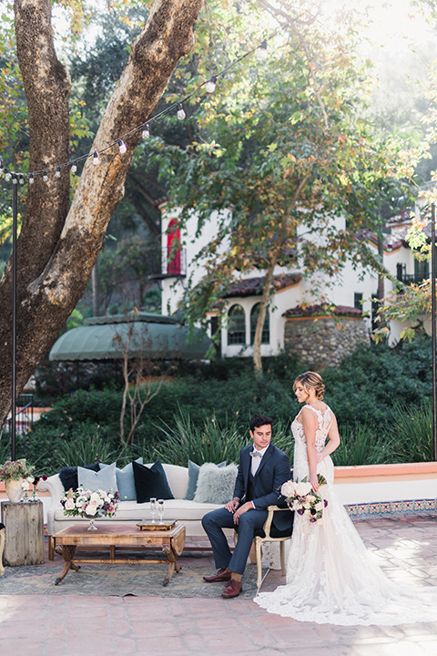 rancho-las-lomas-wedding-groom-sitting-bride-with-bouquet-behind-her-back-bride-wearing-a-dress-with-an-illusion-detail-and-straps-with-lace-on-the-dress-groom-wearing-a-dark-blue-suit-with-a-grey-velvet-bow-tie