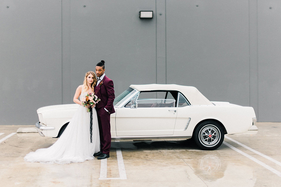 rock-n-roll-wedding-style-bride-and-groom-in-front-of-car-bride-in-a-white-flowing-gown-and-a-black-leather-jacket-groom-in-a-deep-burgundy-suit-with-floral-bow-tie