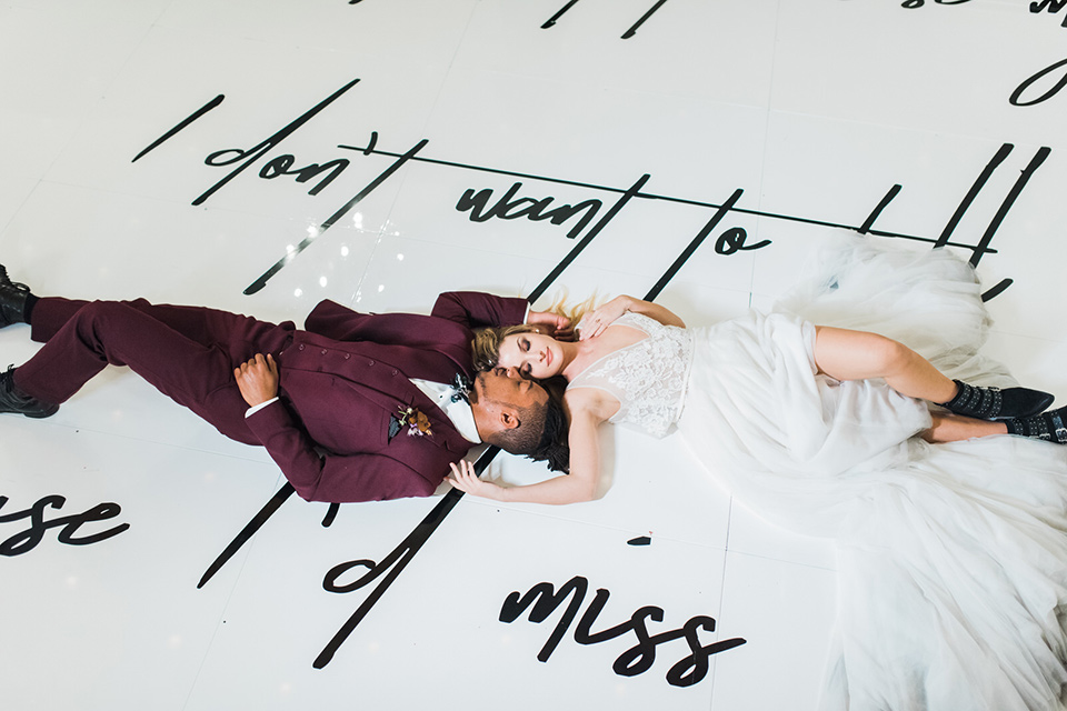 rock-n-roll-wedding-style-bride-and-groom-laying-on-ground-bride-in-a-white-flowing-gown-and-a-black-leather-jacket-groom-in-a-deep-burgundy-suit-with-floral-bow-tie