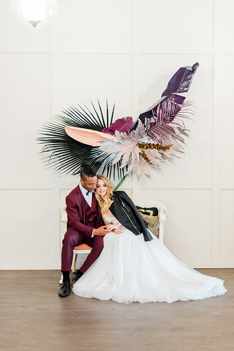 rock-n-roll-wedding-style-bride-and-groom-sitting-on-white-couch-bride-in-a-white-flowing-gown-and-a-black-leather-jacket-groom-in-a-deep-burgundy-suit-with-floral-bow-tie