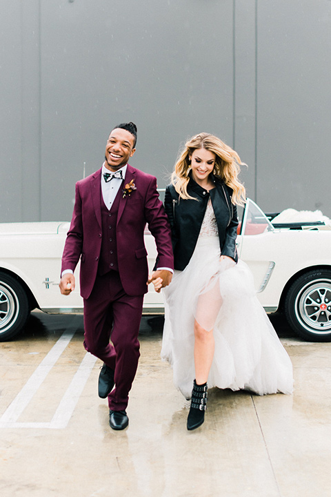 rock-n-roll-wedding-style-bride-and-groom-walking-bride-in-a-white-flowing-gown-and-a-black-leather-jacket-groom-in-a-deep-burgundy-suit-with-floral-bow-tie