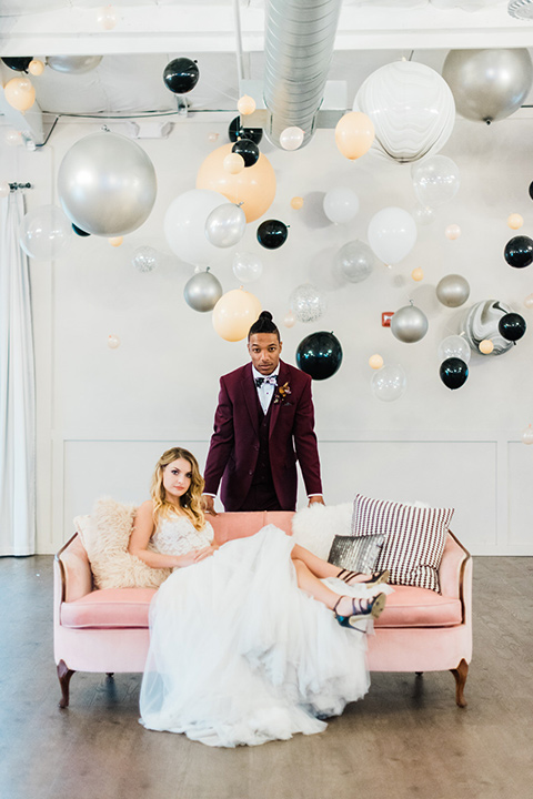 rock-n-roll-wedding-style-bride-on-couch-groom-standing-bride-in-a-white-flowing-gown-and-a-black-leather-jacket-groom-in-a-deep-burgundy-suit-with-floral-bow-tie