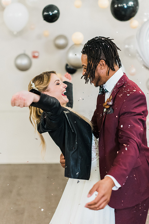 rock-n-roll-wedding-style-bride-with-her-hands-up-groom-smiling-bride-in-a-white-flowing-gown-and-a-black-leather-jacket-groom-in-a-deep-burgundy-suit-with-floral-bow-tie