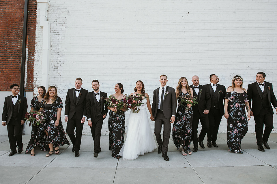 Moniker-Warehouse-Wedding-bridal-party-walking-bridesmaids-in-black-with-pink-floral-long-gowns-the-groomsmen-are-in-black-tuxedos-the-bride-is-in-a-flowing-gown-with-straps-and-a-deep-v-neckline-while-the-groom-wore-a-charcoal-grey-tuxedo-with-a-black-long-tie