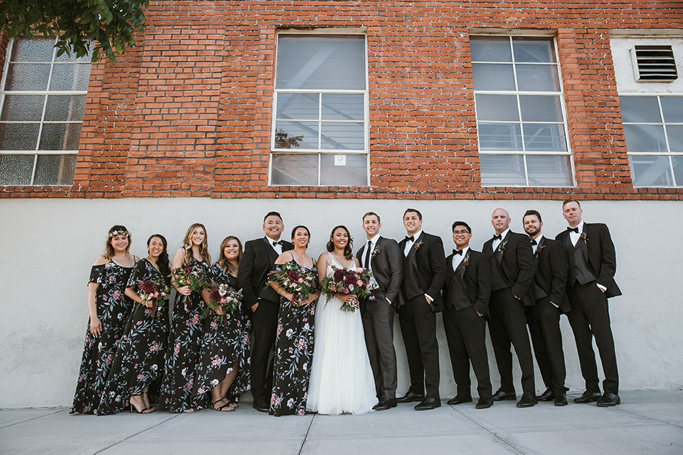 Moniker-Warehouse-Wedding-bridalparty-standing-bridesmaids-in-black-with-pink-floral-long-gowns-the-groomsmen-are-in-black-tuxedos-the-bride-is-in-a-flowing-gown-with-straps-and-a-deep-v-neckline-while-the-groom-wore-a-charcoal-grey-tuxedo-with-a-black-long-tie