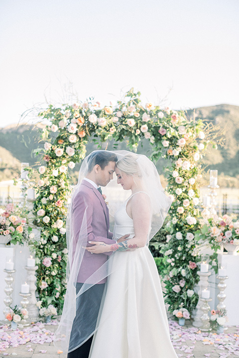  bride in a white ballgown with straps and a deep v neckline and the groom in a pink suit coat with black pants and green velvet bow tie 
