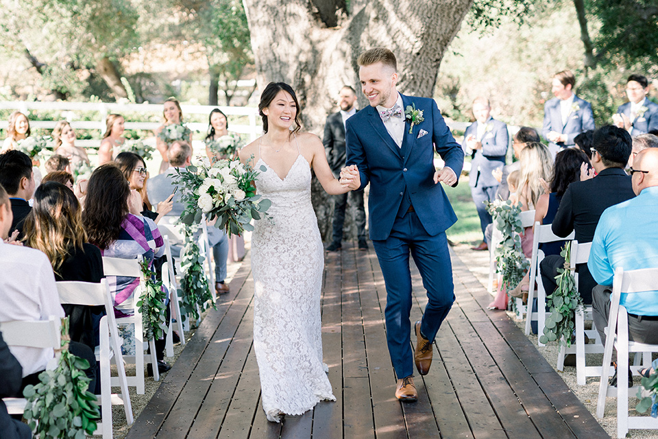 Circle-Oak-ranch-wedding-bride-and-groom-walking-down-aisle-bride-in-lace-form-fitting-gown-with-thin-straps-and-groom-was-in-a-cobalt-suit-with-a-floral-bowtie