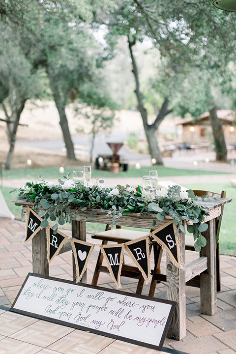 Circle-Oak-ranch-wedding-sweetheart-table-with-a-wooden-table-and-chair-with-greenery-draped-on-it