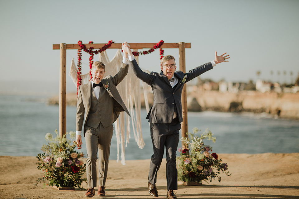 Sunset-Cliffs-Shoot-grooms-at-ceremony-space-one-groom-in-a-grey-suit-with-the-other-groom-in-a-blue-suit-over-looking-the-ocean-with-a-wooden-arch-and-hanging-flowers