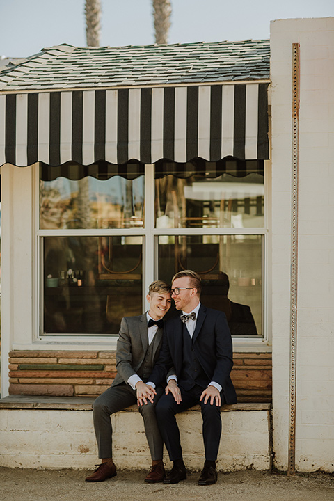 Sunset-Cliffs-Shoot-grooms-sitting-outside-venue-one-groom-in-a-grey-suit-and-the-other-groom-in-a-blue-suit