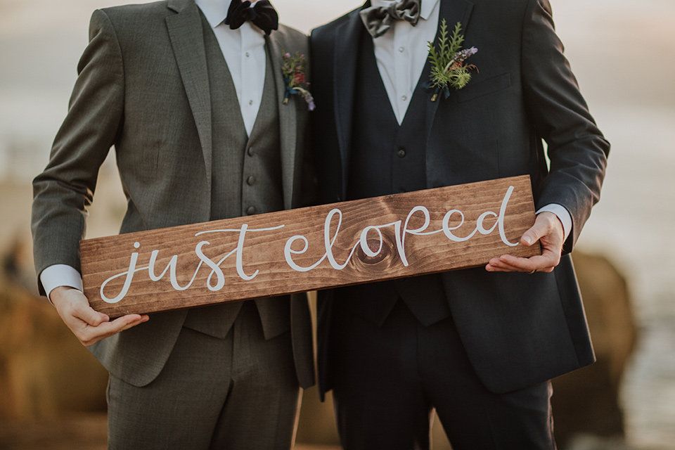 Sunset-Cliffs-Shoot-we-eloped-sign-one-groom-in-a-grey-suit-with-the-other-groom-in-a-blue-suit