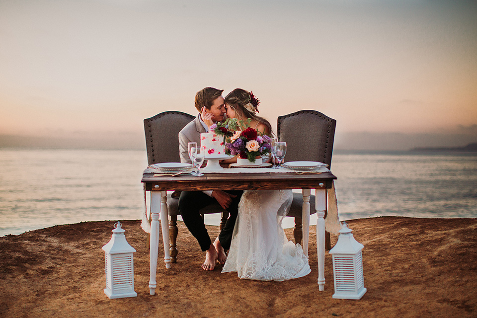 Sunset-Cliffs-Elopement-bride-and-groom-at-sweetheart-table-bride-in-a-lace-form-fitting-gown-with-thin-straps-groom-in-a-tan-suit-coat-with-black-pants-and-black-bow-tie