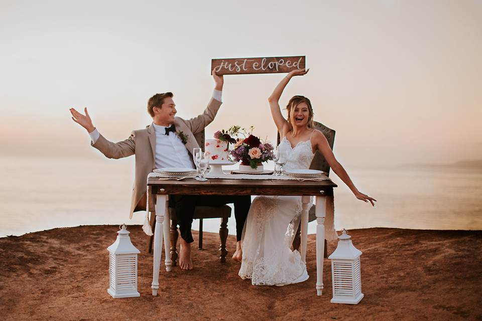 Sunset-Cliffs-Elopement-bride-and-groom-holding-up-elopement-sign-bride-in-a-lace-form-fitting-gown-with-thin-straps-groom-in-a-tan-suit-coat-with-black-pants-and-black-bow-tie