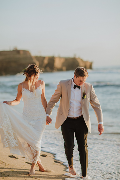 Sunset-Cliffs-Elopement-bride-and-groom-walking-by-water-bride-in-a-lace-white-gown-with-thin-straps-groom-in-a-tan-suit-coat-with-black-pants-and-black-bow-tie