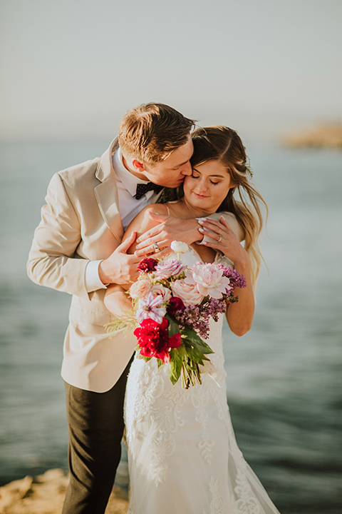 Sunset-Cliffs-Elopement-groom-hugging-bride-from-behind-bride-in-a-lace-white-gown-with-thin-straps-groom-in-a-tan-suit-coat-with-black-pants-and-black-bow-tie