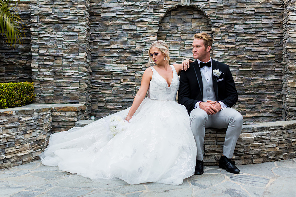 Wedding bride and groom sitting by bricks at the Belamar Hotel in a white ballgown with a deep v neckline and hair in a braid with groom in a black framed tuxedo jacket with a light grey pant
