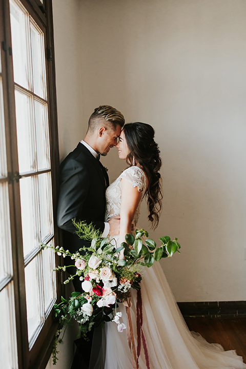 bride in a lace gown with a cap sleeve and the groom in a black suit with a black long tie by the windows