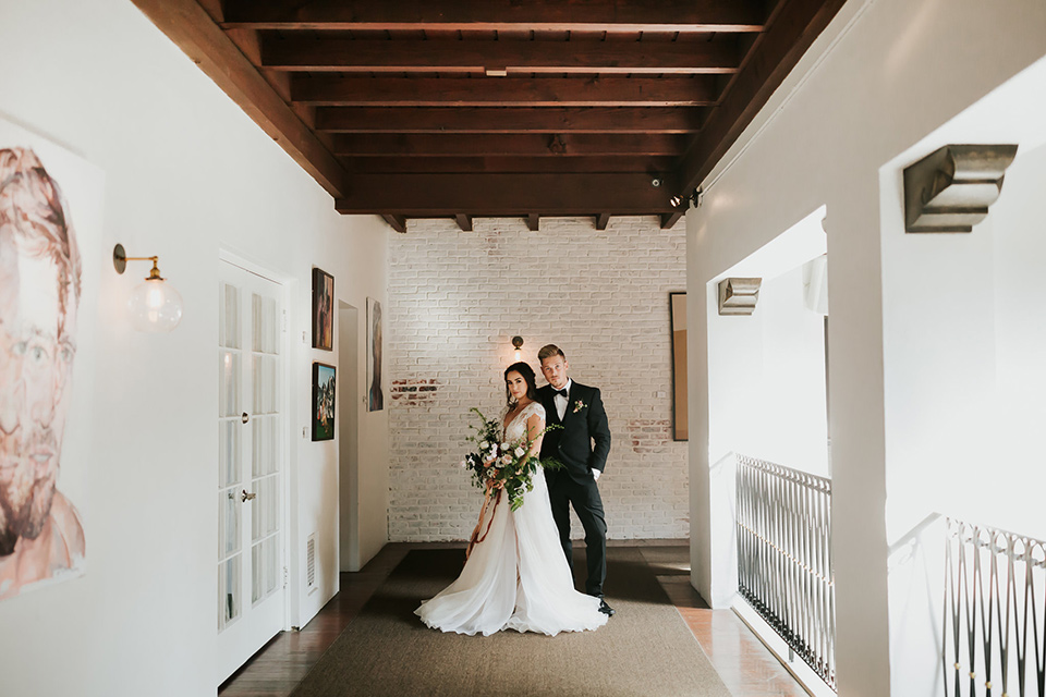 bride in a lace full gown with a cap sleeve and illusion neckline and the groom in a black suit with black long tie standing in hallway