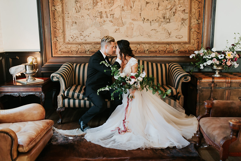 bride in a lace full gown with a cap sleeve and illusion neckline and the groom in a black suit with black long tie sitting on the couch