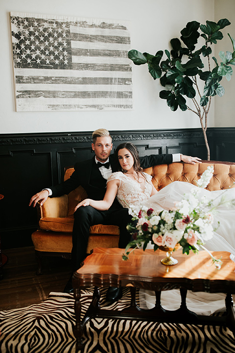bride in a lace gown with a cap sleeve and illusion neckline and the groom in a black suit with a long black tie sitting down on a coach