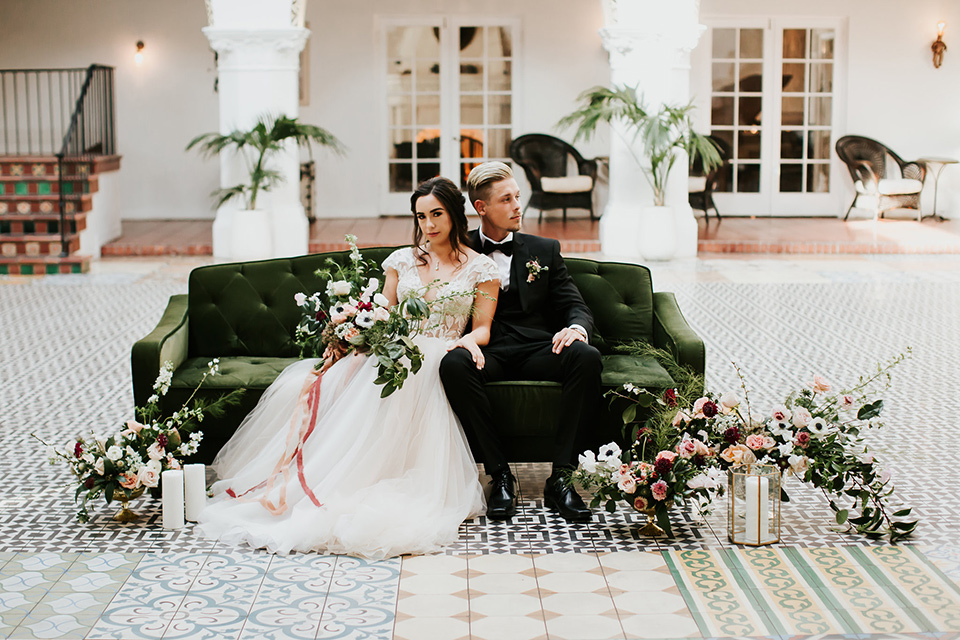 bride in a lace full gown with a cap sleeve and illusion neckline and the groom in a black suit with black long tie sitting on the couch outside