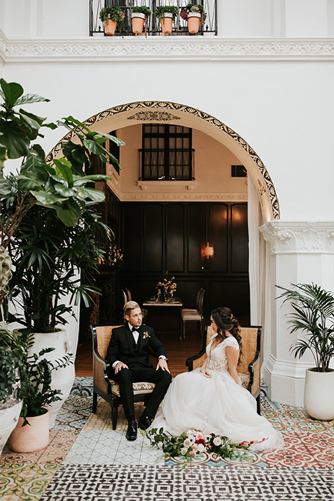 bride in a lace gown with a cap sleeve and the groom in a black suit with a black long tie sitting on chairs at the venue