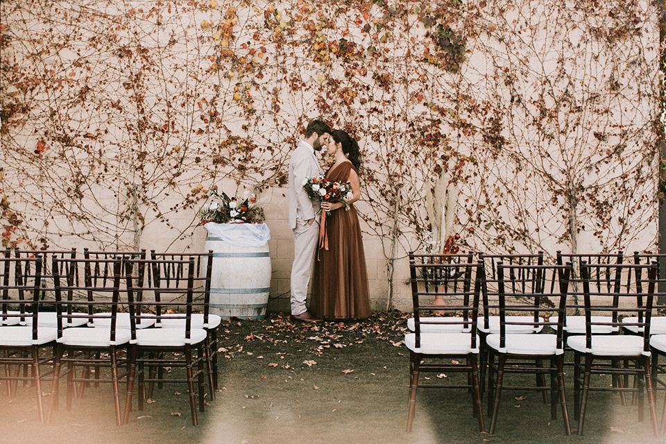  the bride in a chocolate and tan brown gown with a high neckline and sleeves, along with nude suede heels and her hair in a high ponytail and the groom in a tan suit with a chocolate brown long tie