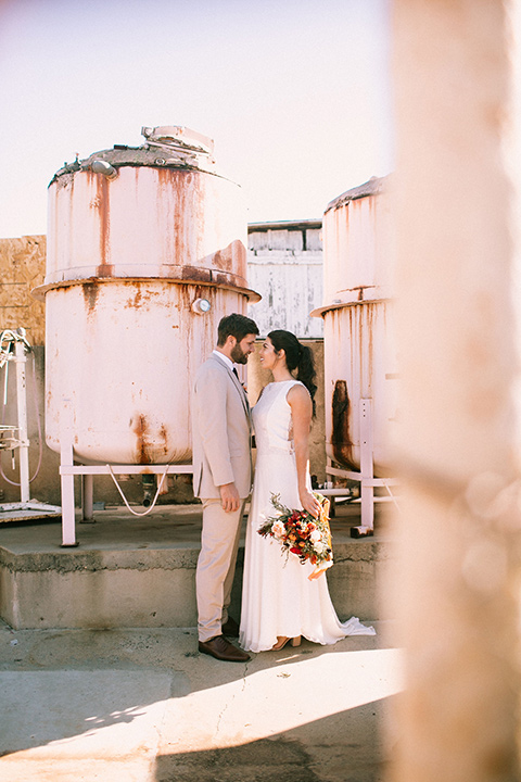 the bride in a white gown with a high neckline and sleeves, along with nude suede heels and her hair in a high ponytail the groom in a tan suit with a chocolate brown long tie