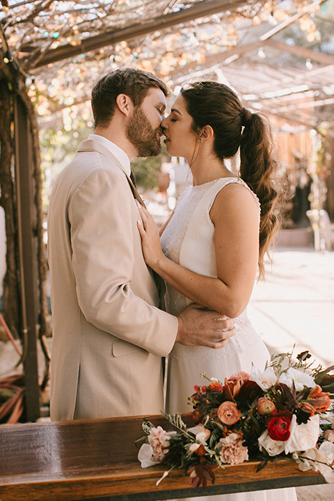  the bride in a white gown with a high neckline and sleeves, along with nude suede heels and her hair in a high ponytail the groom in a tan suit with a chocolate brown long tie 