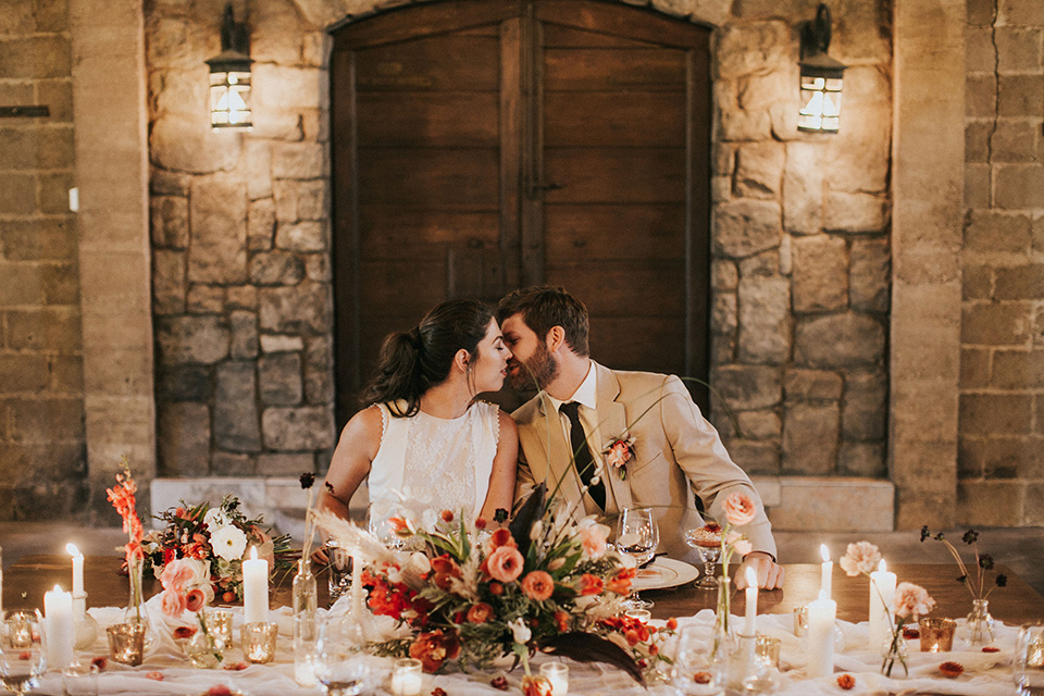  the bride in a white gown with a high neckline and sleeves, along with nude suede heels and her hair in a high ponytail and the groom in a tan suit with a chocolate brown long tie