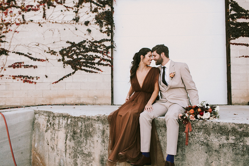 the bride in a chocolate and tan brown gown with a high neckline and sleeves, along with nude suede heels and her hair in a high ponytail and the groom in a tan suit with a chocolate brown long tie