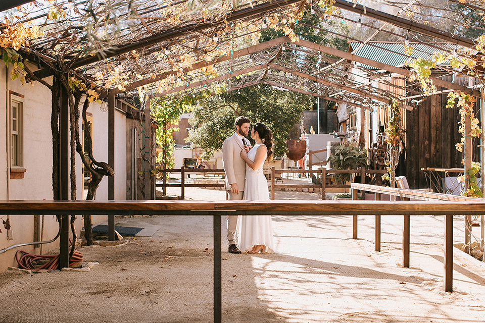  the bride in a white gown with a high neckline and sleeves, along with nude suede heels and her hair in a high ponytail and the groom in a tan suit with a chocolate brown long tie