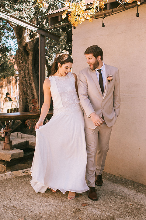 the bride in a white gown with a high neckline and sleeves, along with nude suede heels and her hair in a high ponytail the groom in a tan suit with a chocolate brown long tie