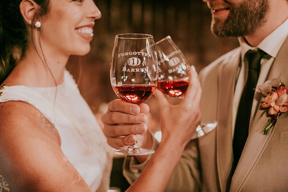  the bride in a white gown with a high neckline and sleeves, along with nude suede heels and her hair in a high ponytail and the groom in a tan suit with a chocolate brown long tie