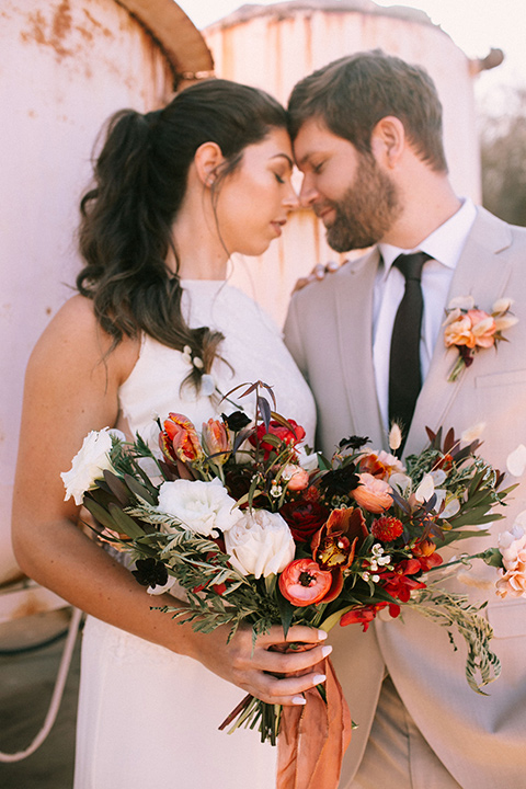  the bride in a white gown with a high neckline and sleeves, along with nude suede heels and her hair in a high ponytail the groom in a tan suit with a chocolate brown long tie 