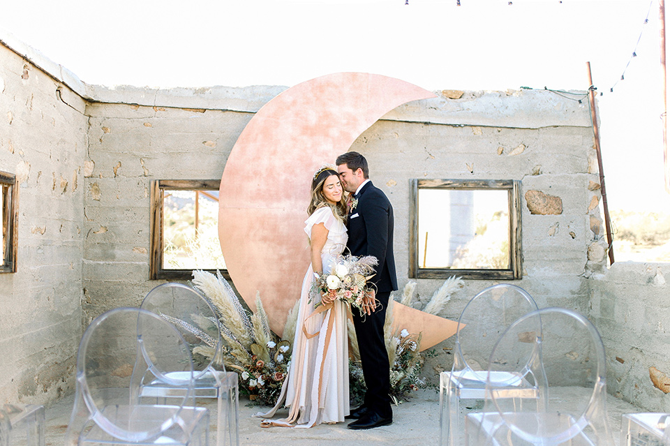 Bride in a cream and ivory flowing gown with a gold headband with stars and the groom in a black tuxedo with a black bow tie by a rose gold moon shaped arch at ceremony