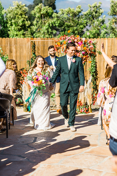 Topa-Winery-Wedding-bride-and-groom-walk-down-aisle-bride-in-a-lace-bohemian-style-gown-with-flutter-sleeves-groom-in-a-green-suit-with-a-tan-long-tie-with-brown-shoes