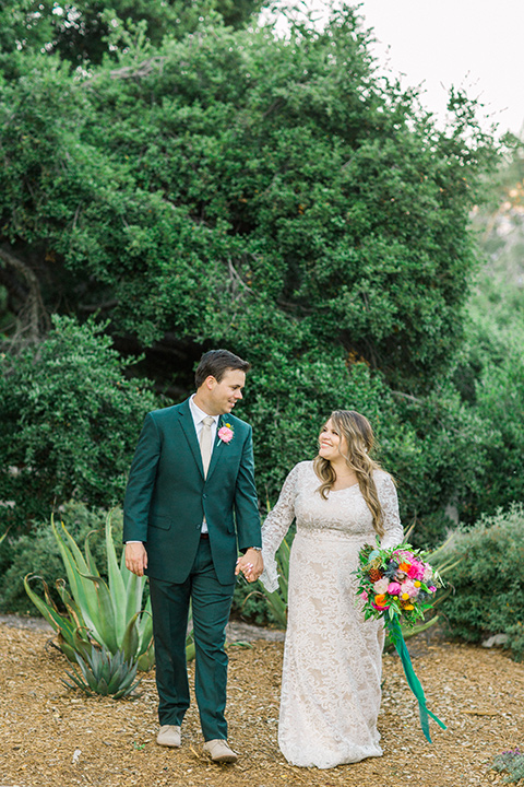 Topa-Winery-Wedding-couple-walking-bride-in-a-bohemian-gown-with-lace-detailing-and-flutter-sleeves-groom-in-a-green-suit-with-a-tan-tie-and-shoes