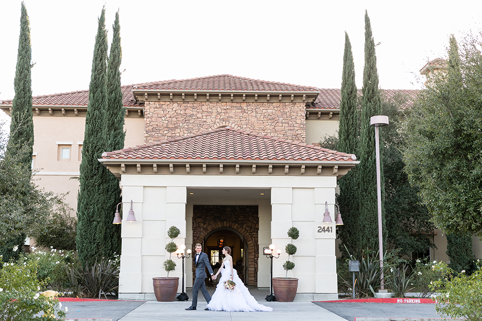 Vellano-Country-Club-bride-and-groom-walking-past-venue-bride-in-a-ballgown-with-her-hair-up-groom-in-a-charcoal-grey-tuxedo-with-a-black-bow-tie