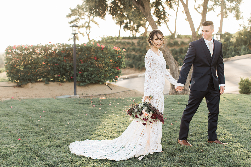 Temecula-outdoor-wedding-at-callaway-winery-bride-and-groom-holding-hands-and-walking-bride-wearing-a-lace-boho-lace-dress-with-sleeves-and-the-groom-in-a-navy-suit-with-champagne-long-tie