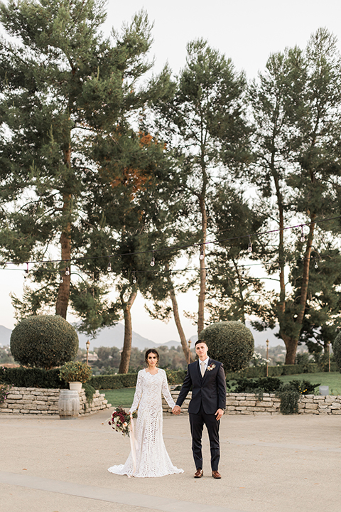 Temecula-outdoor-wedding-at-callaway-winery-bride-and-groom-holding-hands-far-away-with-the-bride-in-a-lace-gown-and-groom-in-a-dark-blue-notch-lapel-suit