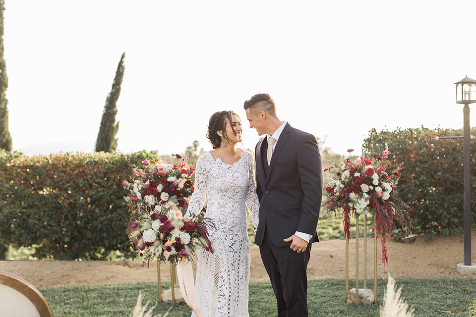 Temecula-outdoor-wedding-at-callaway-winery-bride-and-groom-holding-hands-bride-ina-boho-lace-dress-with-sleeves-and-the-groom-in-a-navy-suit