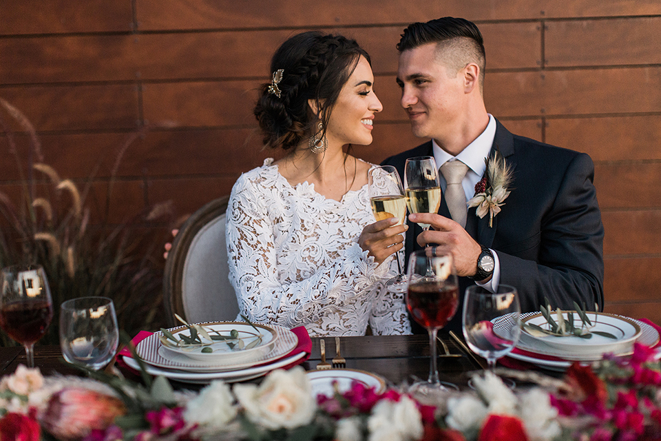 Temecula-outdoor-wedding-at-callaway-winery-table-set-up-sweetheart-table-bride-and-groom-close-up-sitting-with-the-bride-in-a-lace-gown-with-a-high-neckline-and-lace-sleeves-groom-in-a-navy-suit-with-champagne-long-tie