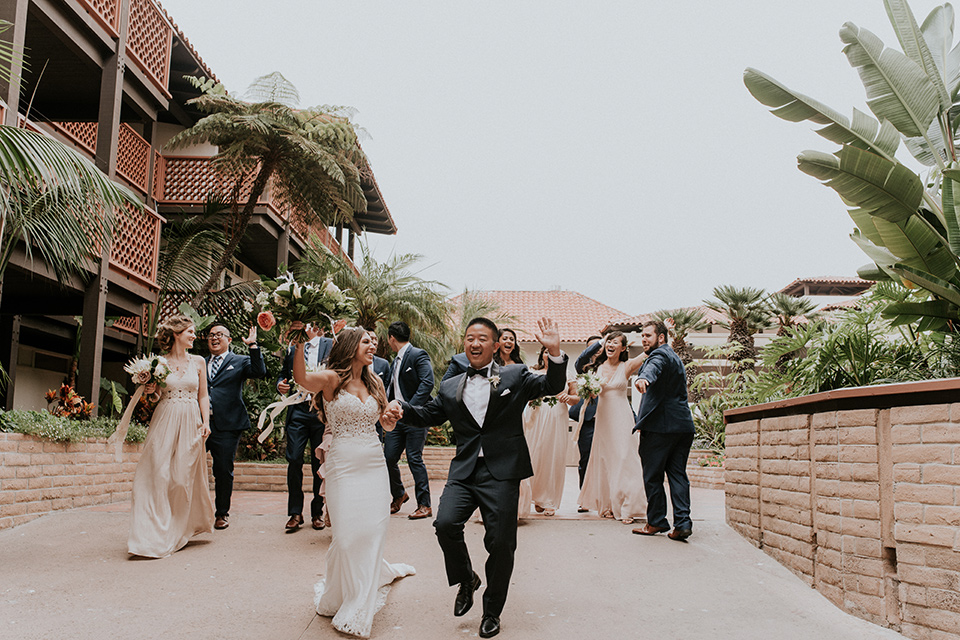 la-jola-shores-hotel-wedding-bridal-party-walking-bridesmaids-in-pink-dresses-groomsmen-in-dark-blue-suits-bride-in-a-formfitting-satin-dress-with-straps-groom-in-a-navy-tuxedo-with-black-shawl-lapel