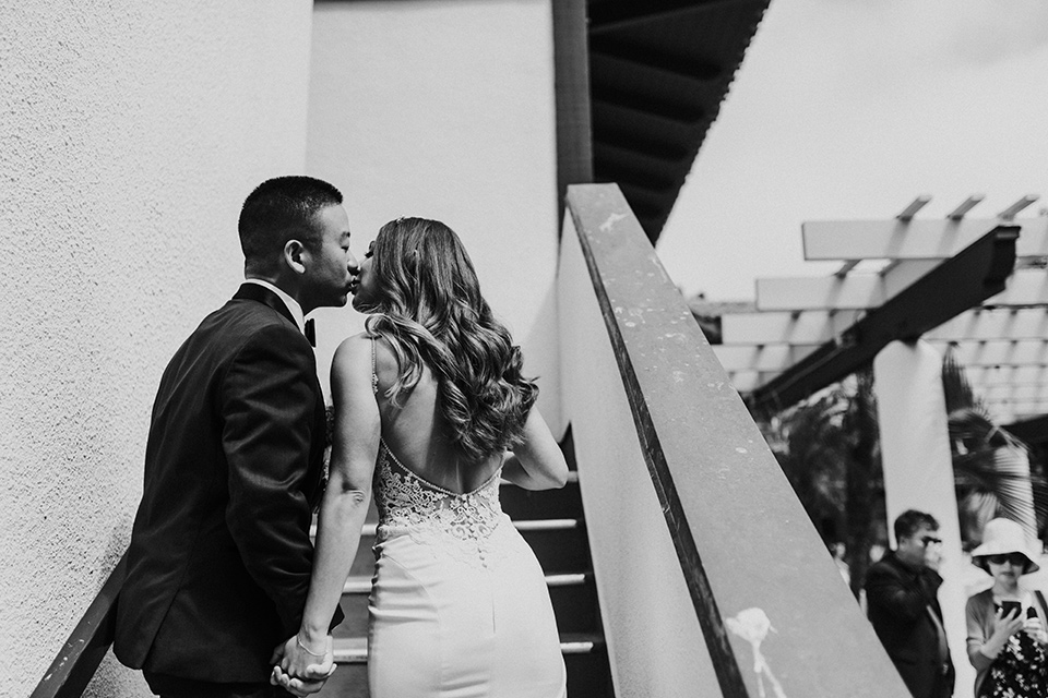 la-jola-shores-hotel-wedding-bride-and-groom-kissing-on-the-stairs-bride-in-a-formfitting-satin-dress-with-straps-groom-in-a-navy-tuxedo-with-black-shawl-lapel