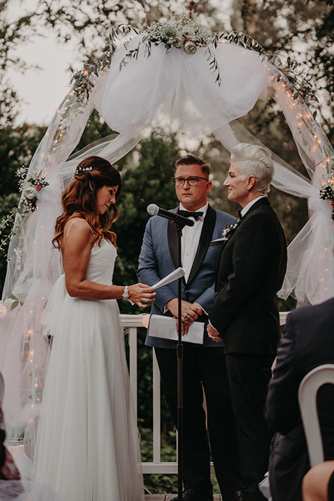 two-brides-at-their-ceremony-one-bride-in-a-flowing-white-gown-the-other-in-a-black-tuxedo-with-a-grey-vest-and-black-bowtie