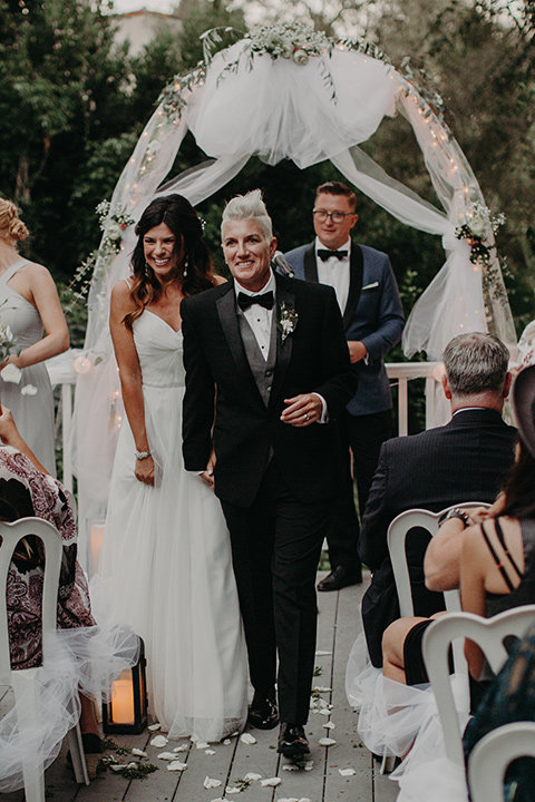 two-brides-at-their-ceremony-one-bride-in-a-flowing-white-gown-the-other-in-a-black-tuxedo-with-a-grey-vest-and-black-bowtie