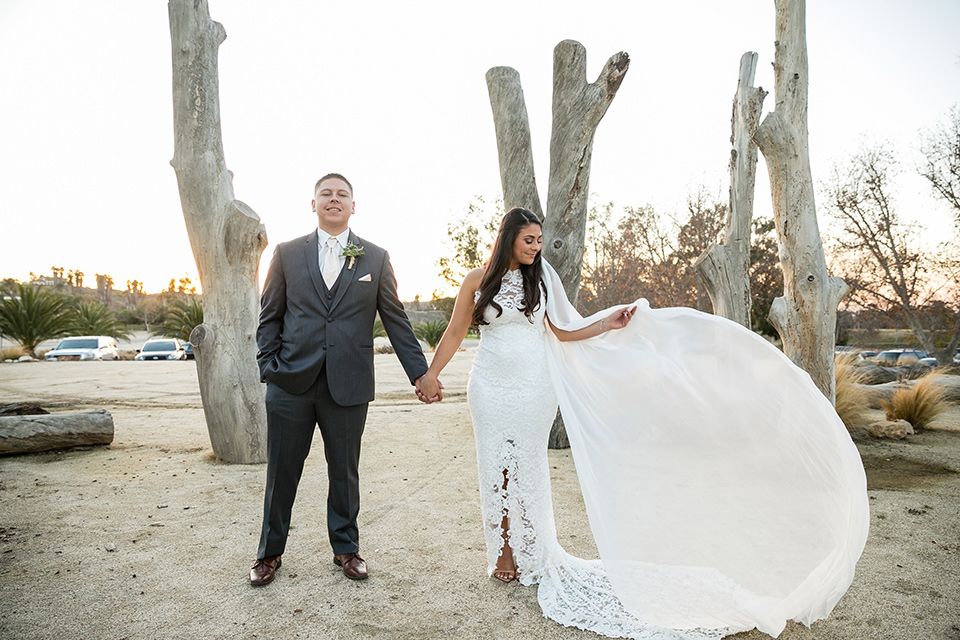 gallway-downs-wedding-bride-and-groom-standing-outside-near-tree-stunps-bride-in-a-lace-form-fitting-gown-with-a-high-neckline-and-keyhole-back-groom-in-a-charcoal-tux-withan-ivory-long-tie