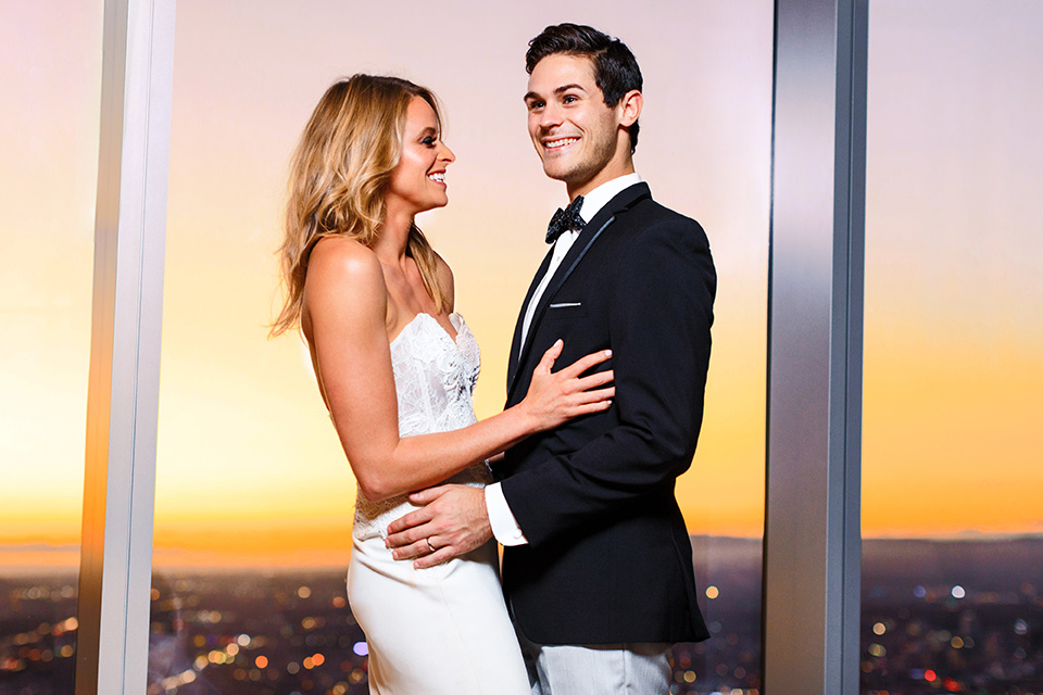 Intercontinental-DTLA-shoot-bride-and-groom-inside-with-sunset-behind-them-laughing-bride-wearing-a-stapless-white-gown-with-lace-detail-and-the-groom-wearing-a-two-toned-tuxedo-in-black-and-grey