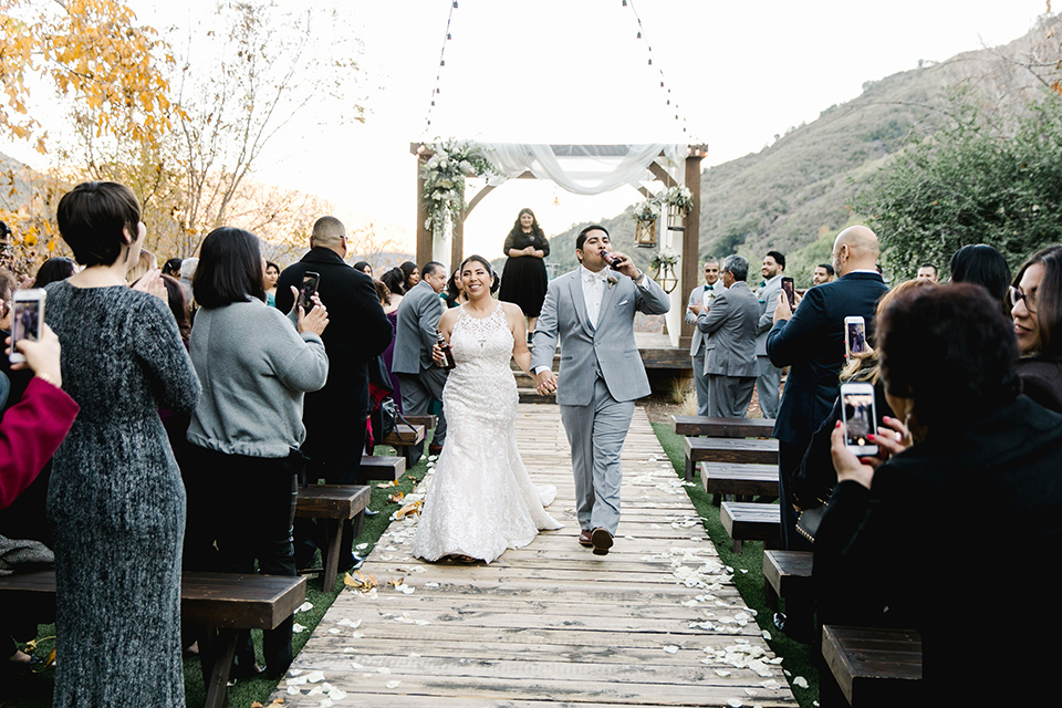 hidden-acres-wedding-drinking-beers-as-they-end-ceremony-bride-in-fitted-gown-with-an-illusion-neckline-and-groom-in-a-grey-suit-with-a-white-bow-tie